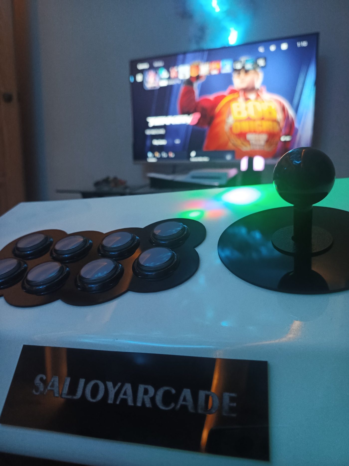 A white SALjoyArcade stick with black buttons and a black ball top, in front of a TV